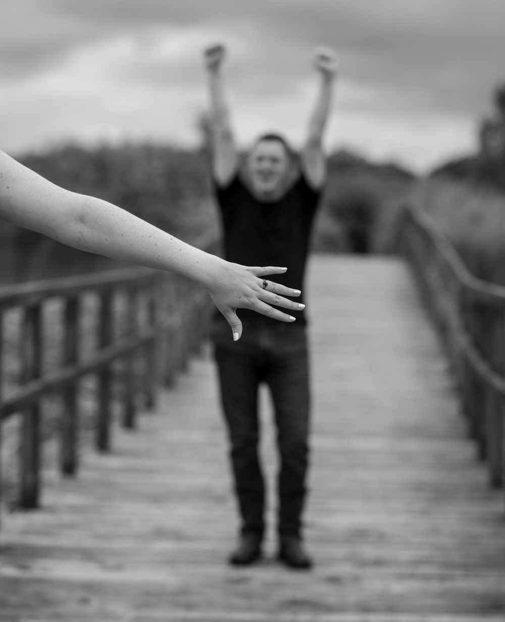 Black and white photo of a hand reaching toward a man celebrating with arms raised on a wooden bridge.