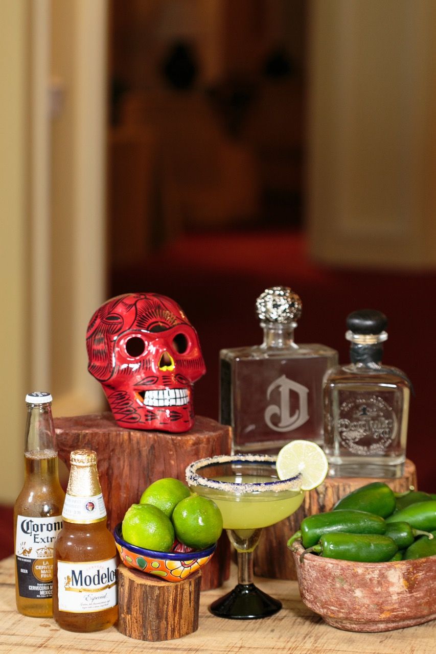 Mexican bar setup with Corona, Modelo, margarita, limes, jalapenos, tequila bottles, and a red Day of the Dead skull.
