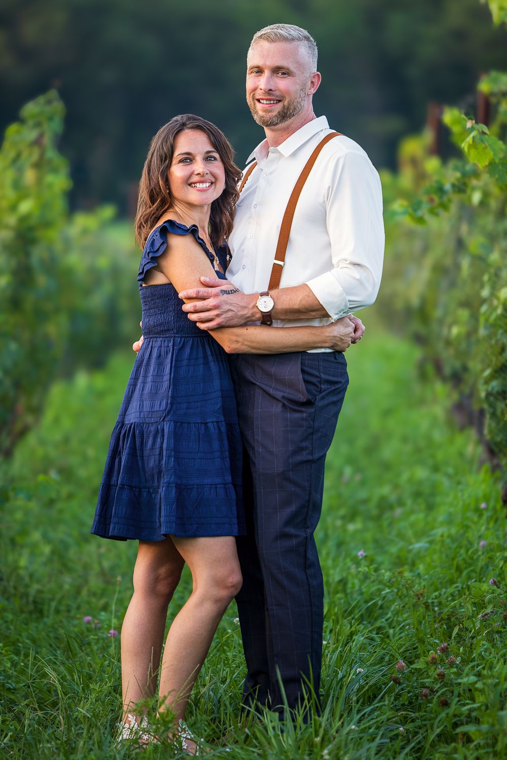 Couple embracing in a summer field wearing navy dress and white shirt with suspenders against natural backdrop.