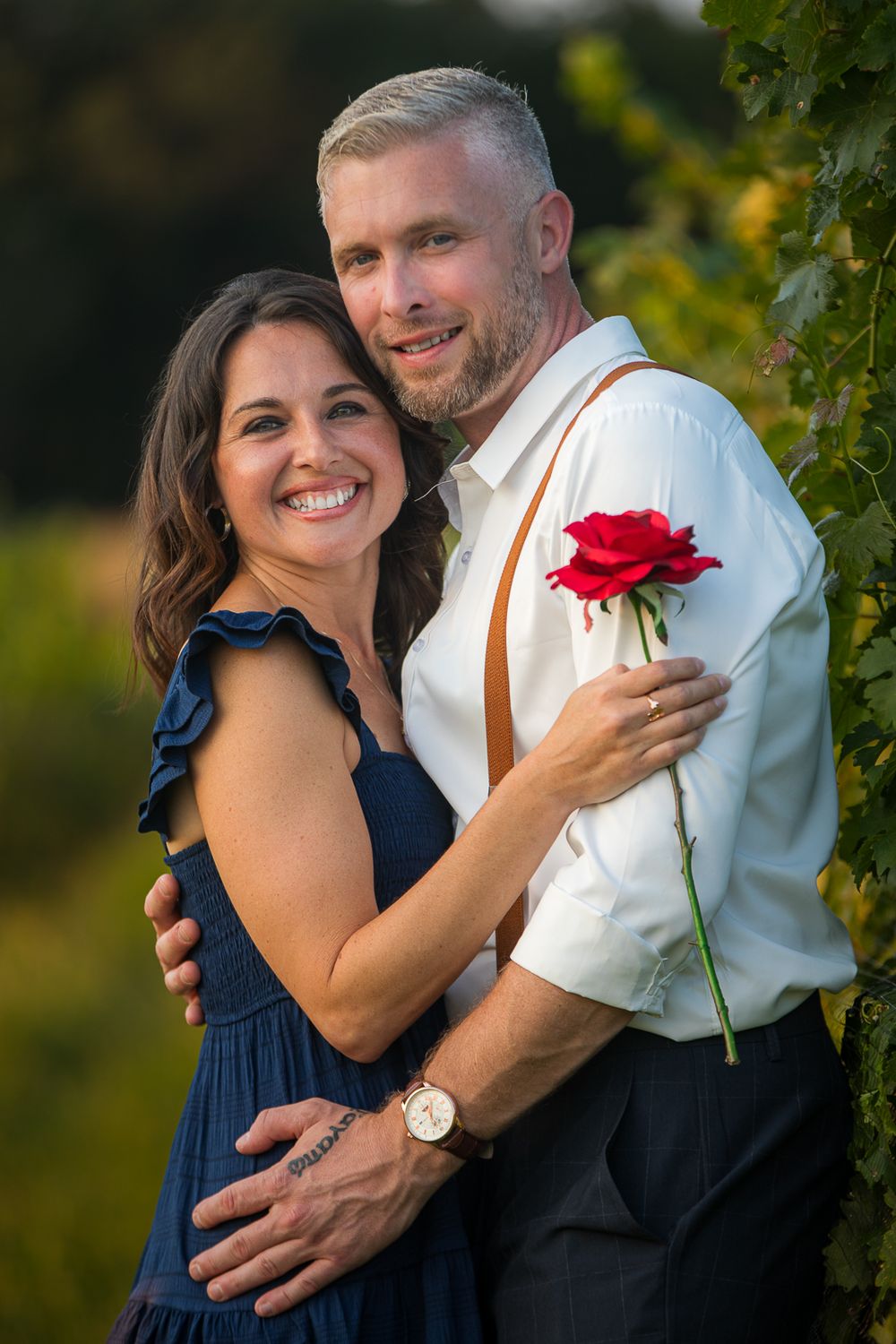 A couple embraces outdoors in formal attire, with a red rose and brown leather suspenders as romantic accessories.