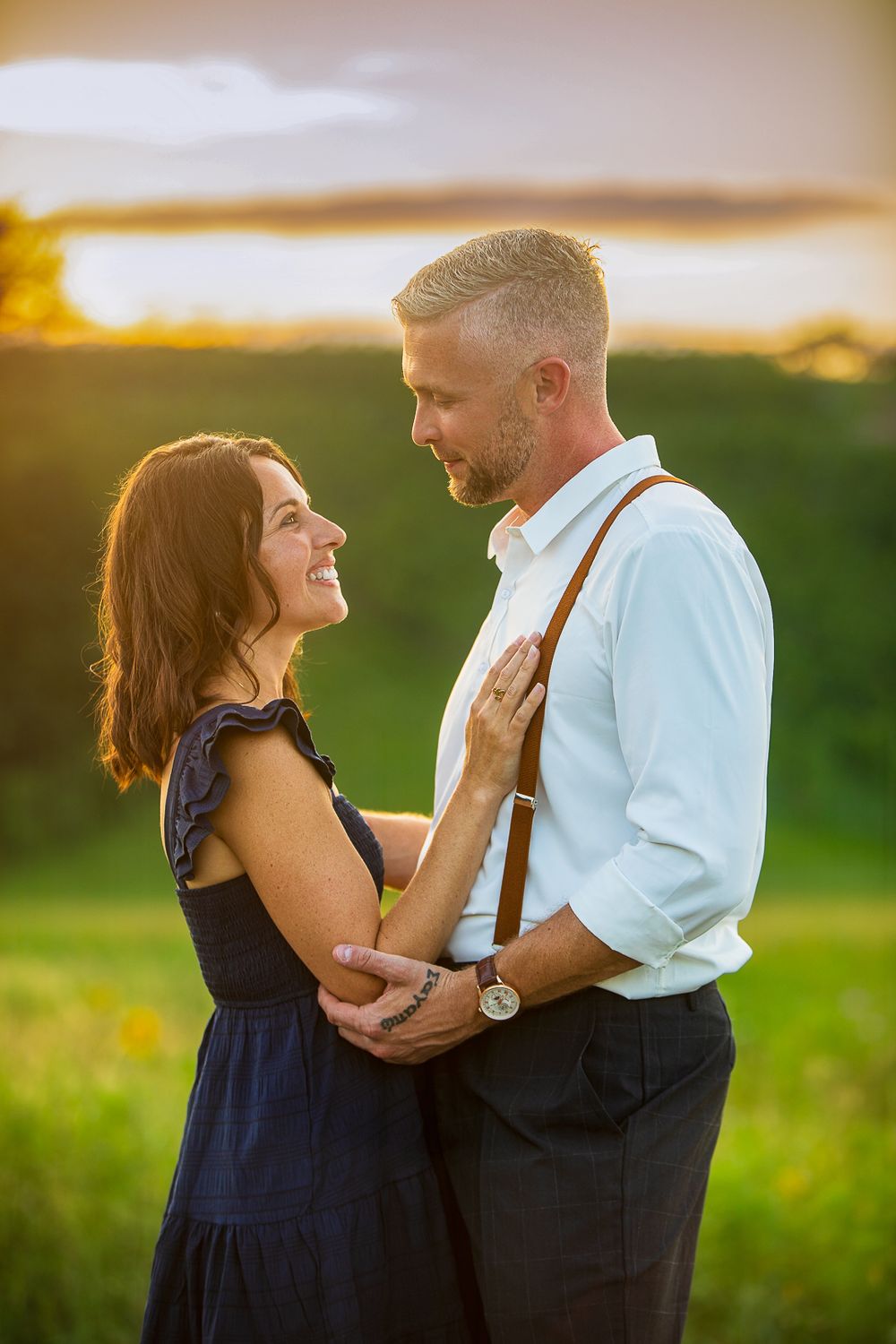 A romantic couple embraces in a field at sunset, with the woman wearing a navy dress and man in suspenders.