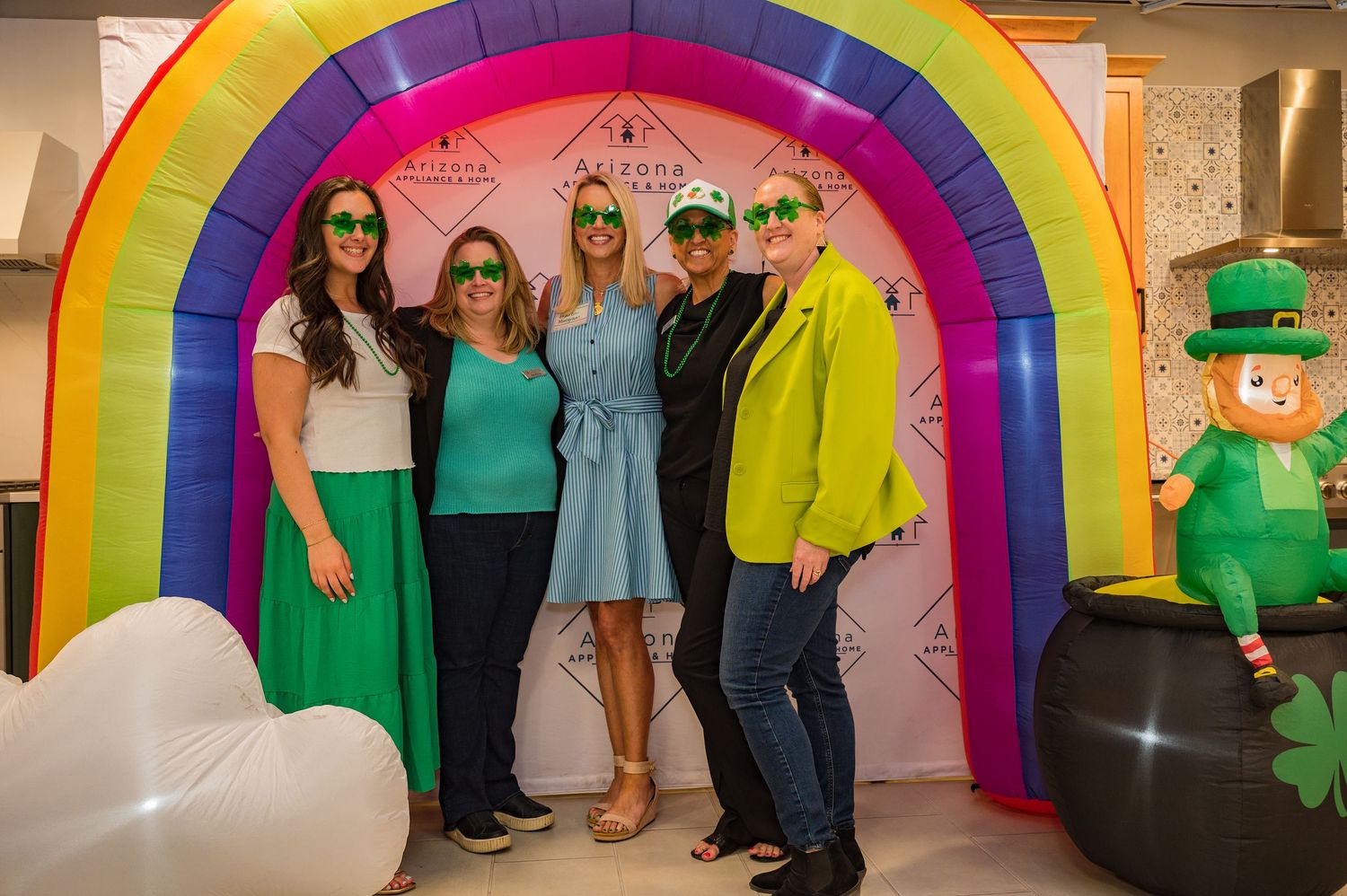 Four guests posing under an inflatable rainbow arch with green sunglasses at a St. Patricks Day party.
