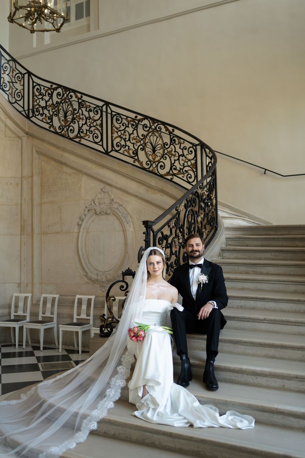 Elegant wedding couple poses on grand marble staircase with ornate wrought iron railing in luxurious venue.