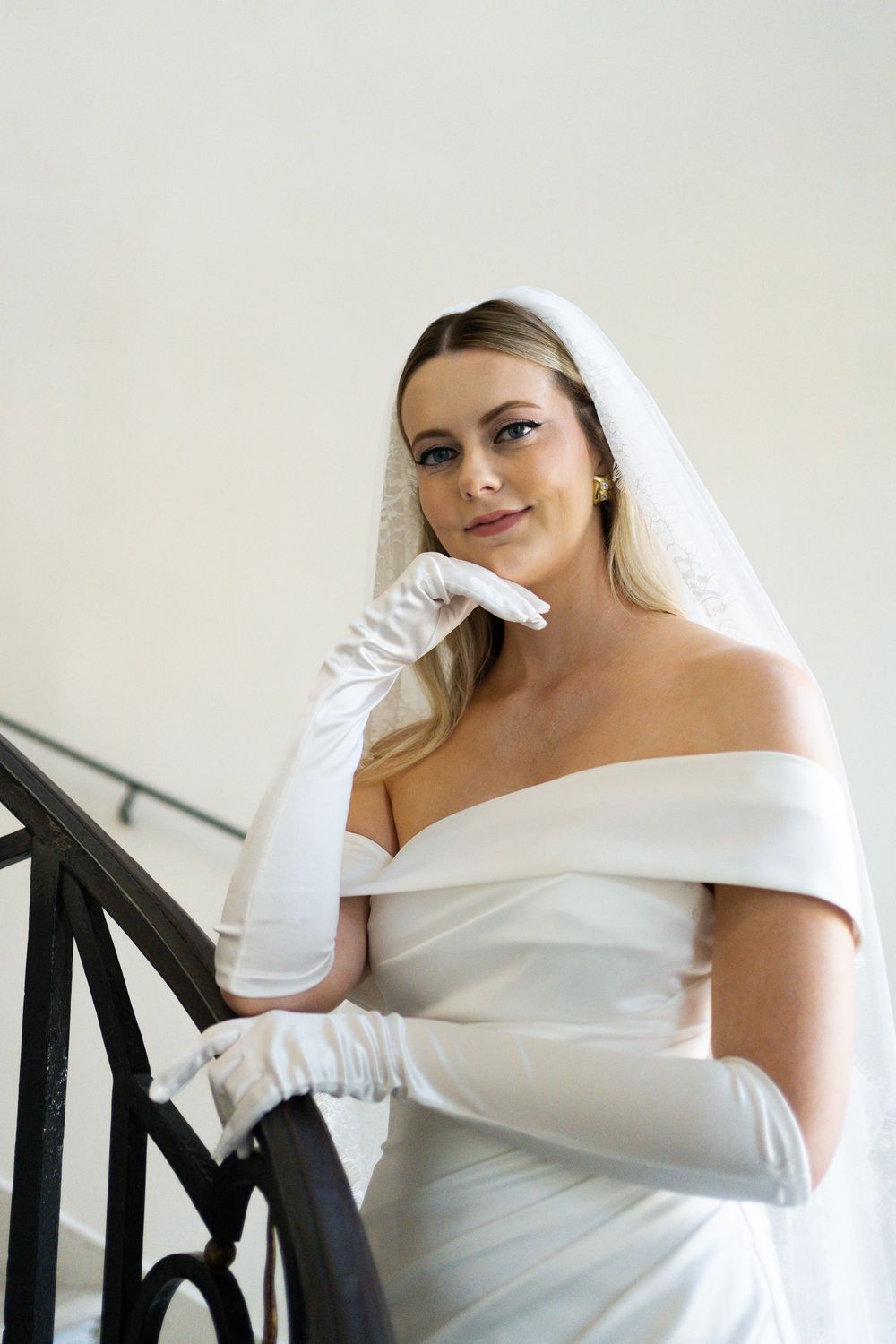 Elegant bride in off-shoulder white wedding gown and long gloves poses gracefully on staircase.