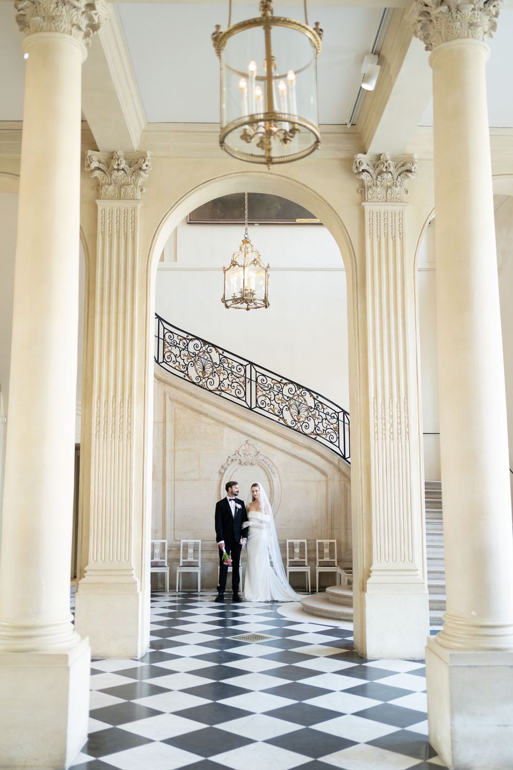 Elegant wedding couple photographed in a grand columned hallway with black and white checkered marble floors and chandeliers.