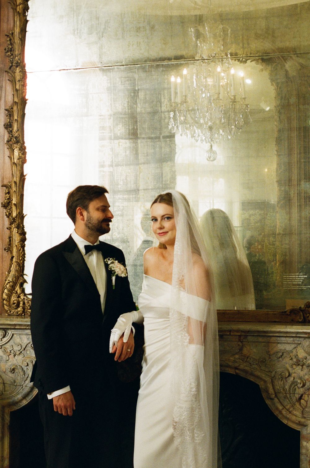 A couple stands in an elegant room with vintage decor and a crystal chandelier overhead.