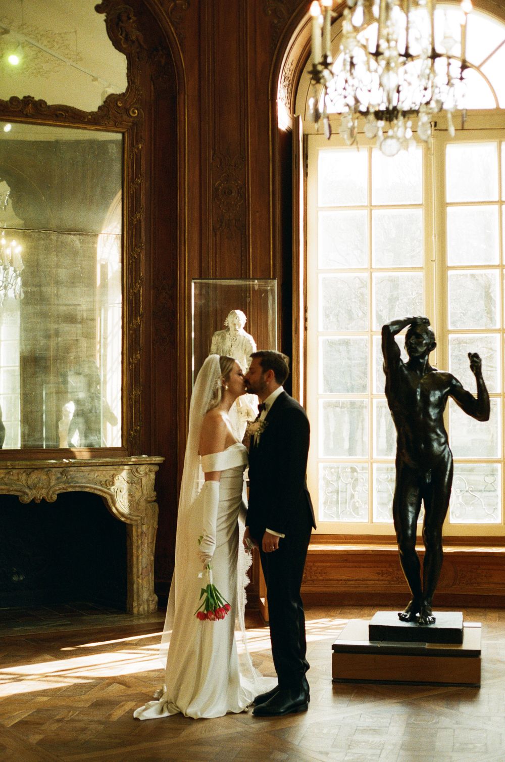 Elegant couple in formal attire embraces in an opulent room with chandelier, fireplace, and statue by large windows.