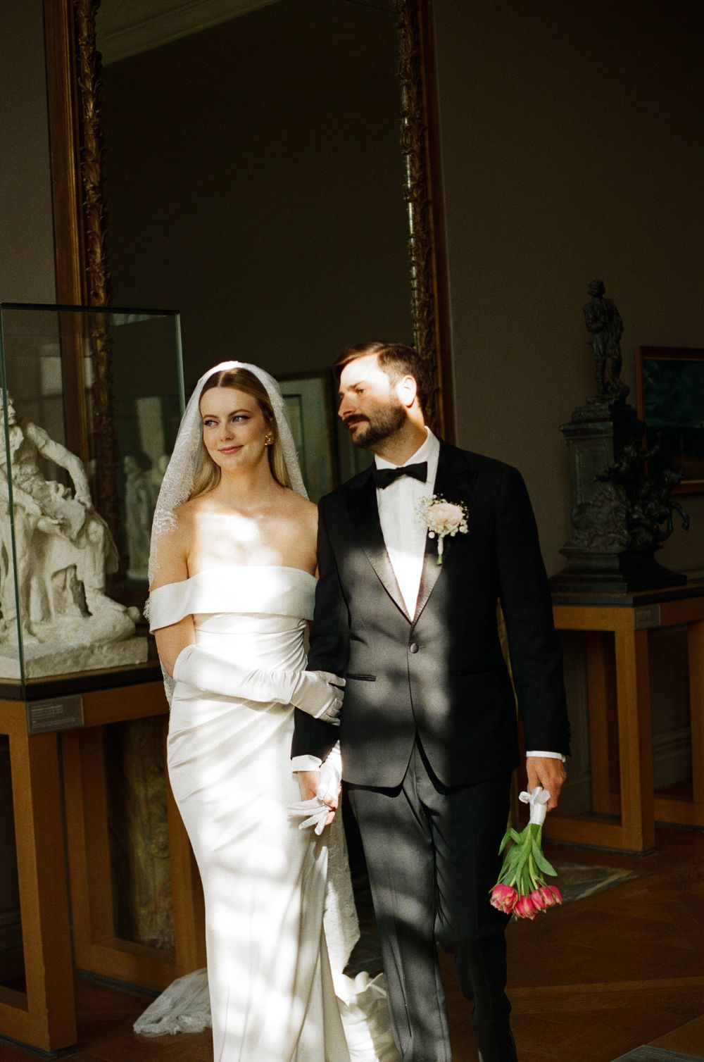 A bride and groom walk together in a dimly lit indoor venue with wooden paneling and artwork on display.