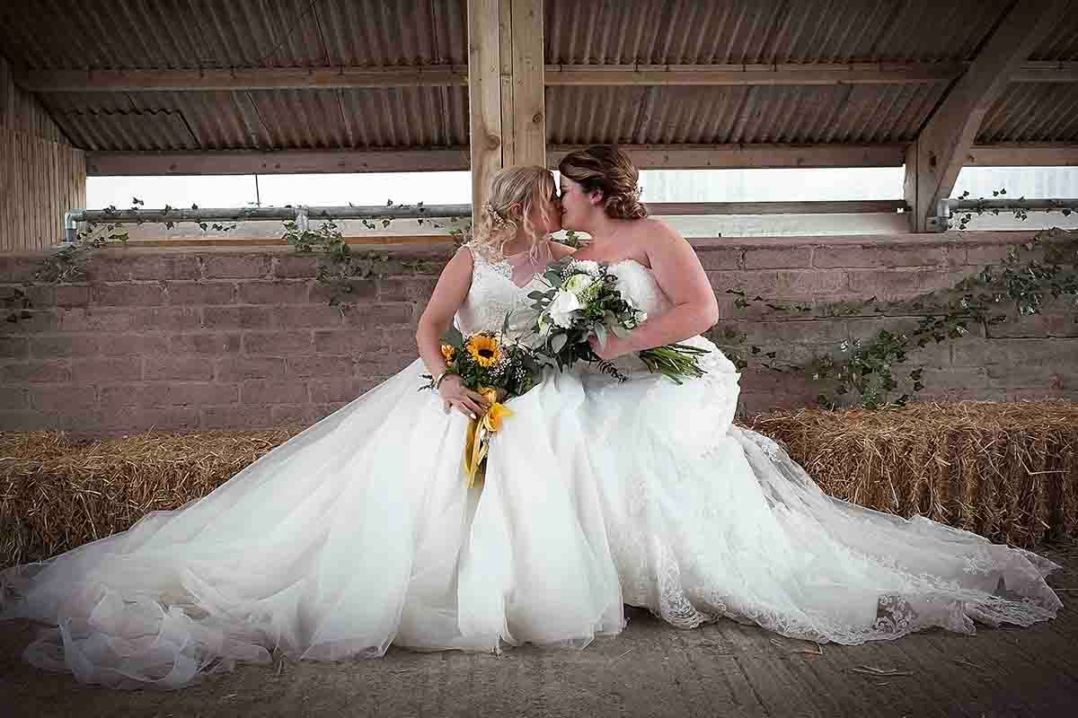 Two brides in white gowns share a romantic moment on hay bales, holding a sunflower bouquet at a rustic barn wedding.