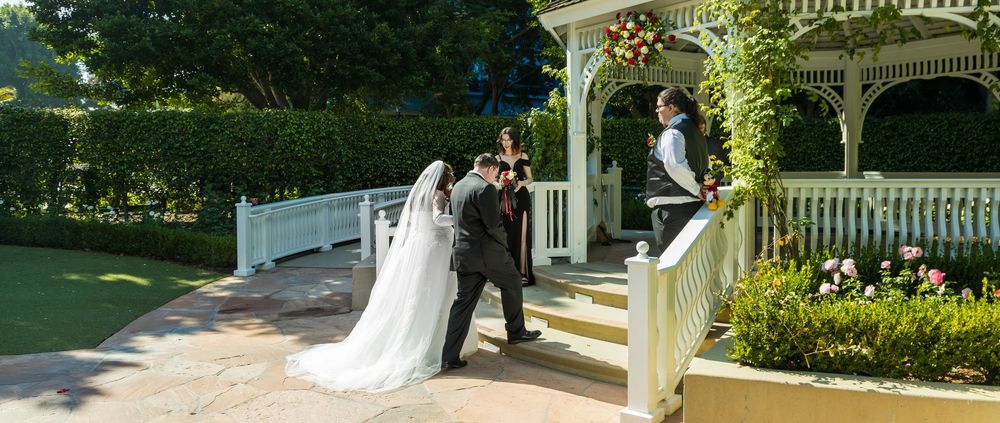 A romantic garden wedding ceremony takes place under an elegant white gazebo surrounded by lush greenery and flowers.