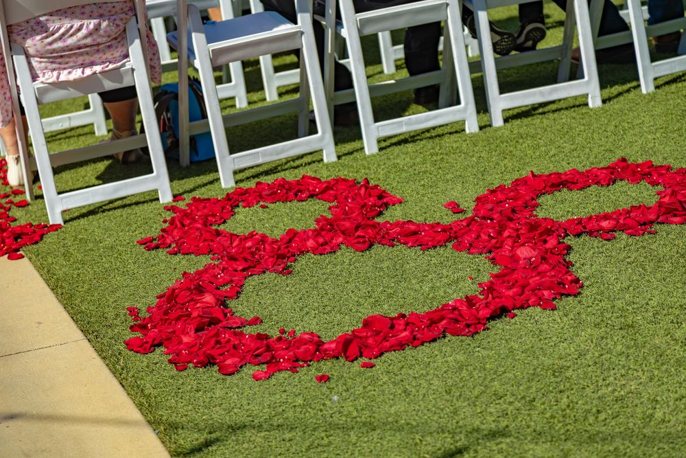 Red rose petals arranged in a heart shape on green grass with white chairs in the background.