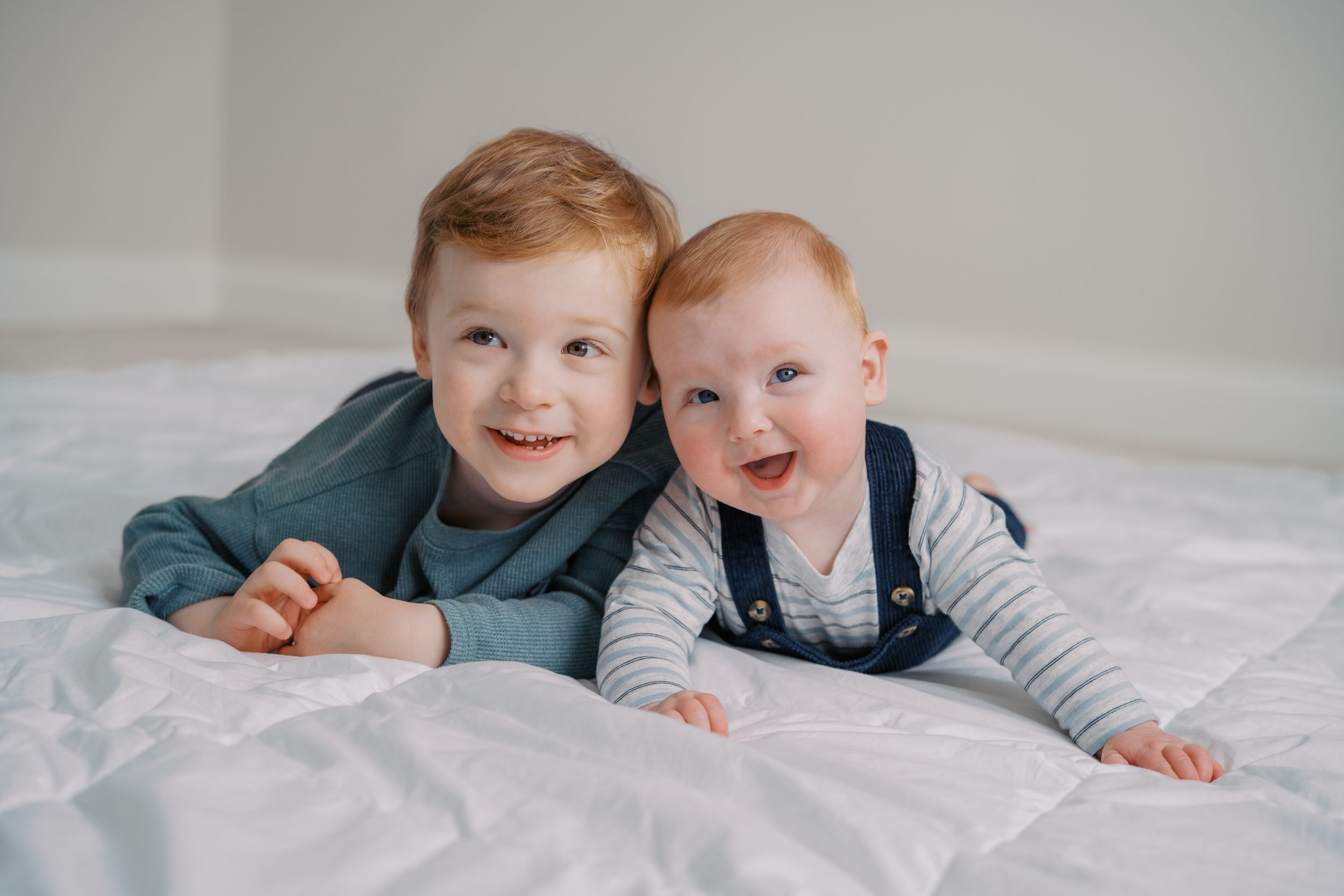Two children lying together on white bedding while playing and laughing.