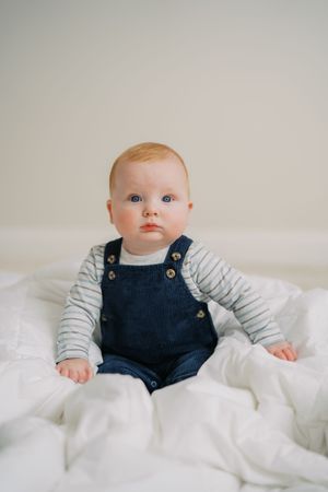 Baby wearing navy overalls and striped shirt sits on white bedding with neutral background.