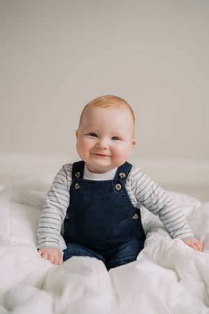 A smiling baby wearing navy corduroy overalls and striped shirt sits on white bedding during a portrait session.