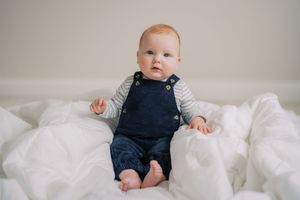 Baby wearing navy blue overalls sits and plays on white bedding while looking away from camera.