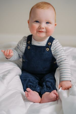Baby wearing navy corduroy overalls and striped shirt sits smiling on white bedding.