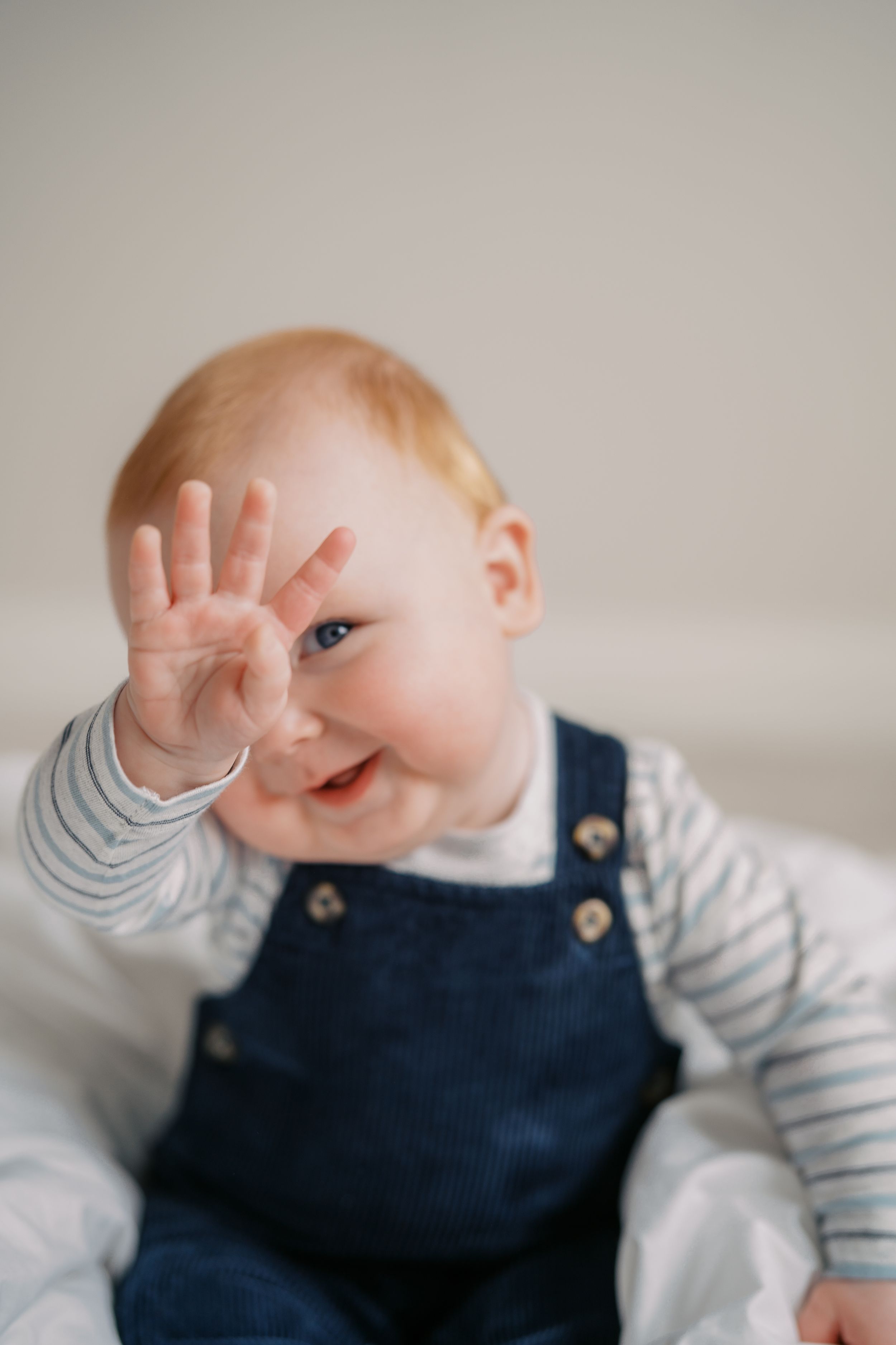 Baby in navy overalls and striped shirt smiling and waving while sitting on light background.
