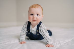 Baby in striped shirt and blue overalls crawls on white bedding in a series of connected photos.