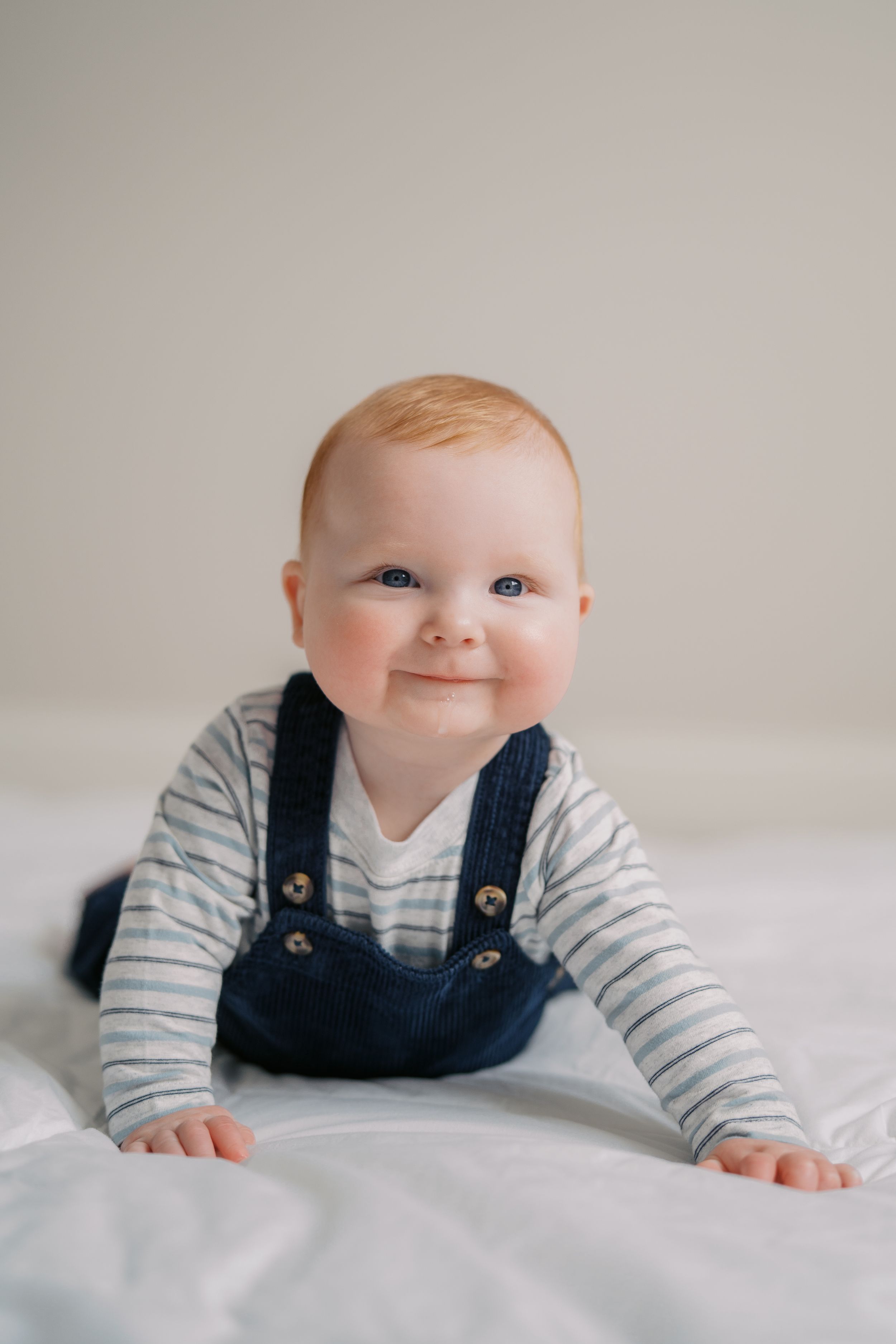Baby in navy overalls and striped shirt smiles while lying on white bedding.
