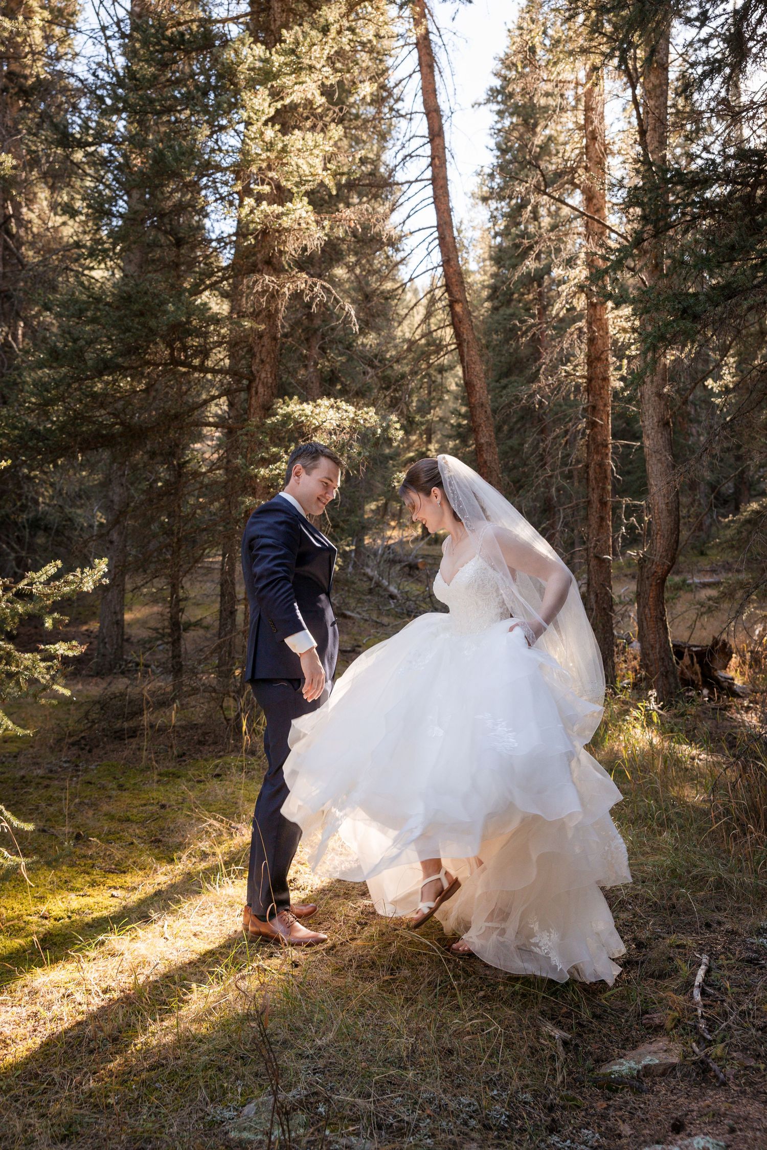 A couple in wedding attire dances together in a sunlit forest setting, creating a romantic outdoor scene.