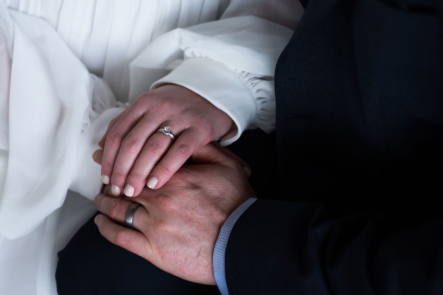 Close-up view of two hands wearing wedding rings against white and black formal attire.