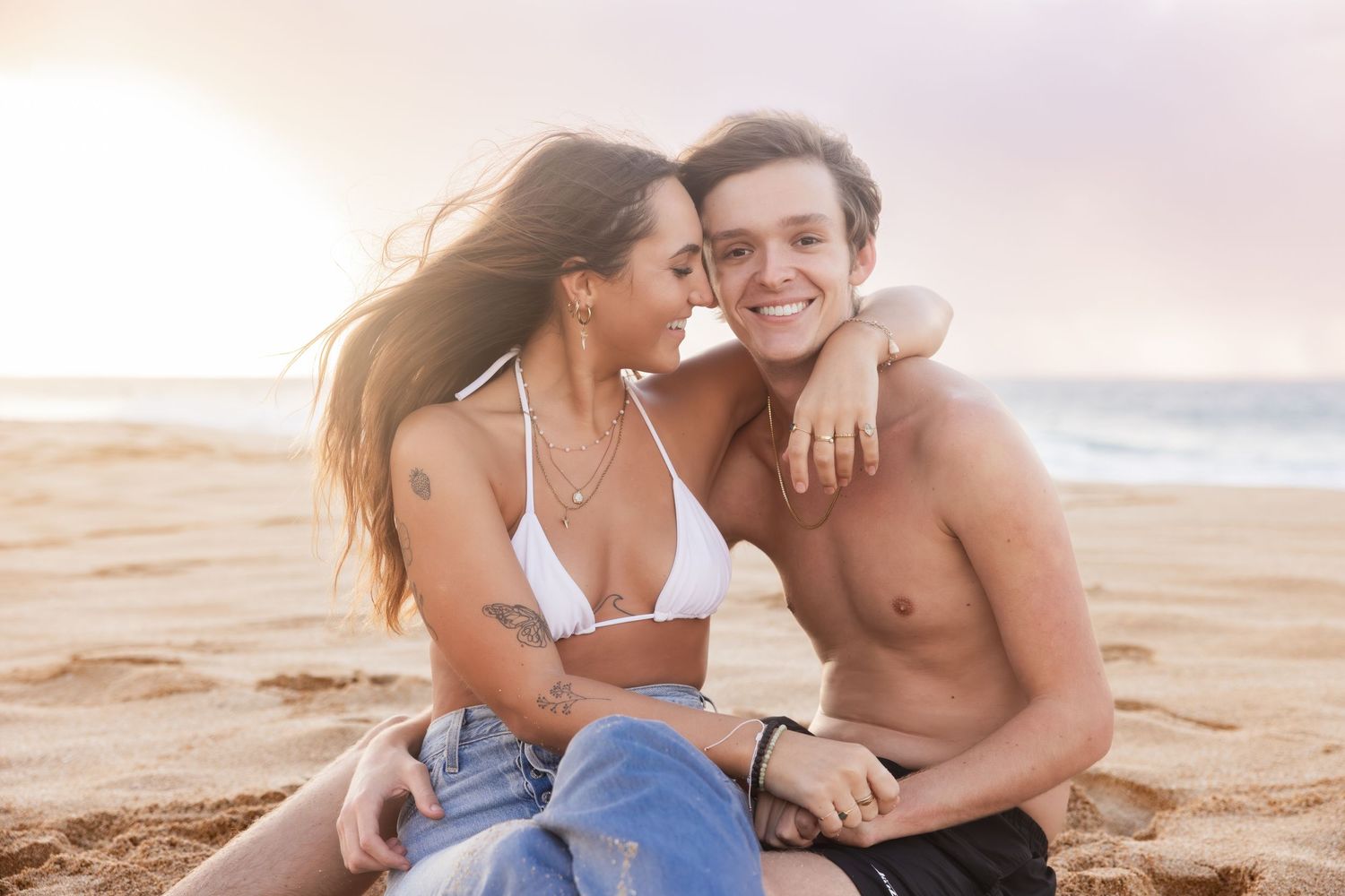 A couple shares intimate moments on a sandy beach at sunset while wearing casual beachwear.