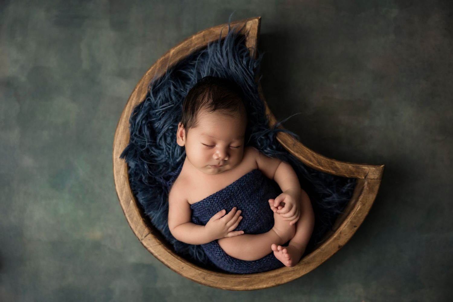 A sleeping newborn baby wearing a navy blue outfit lies peacefully on dark blue fabric during a photography session.