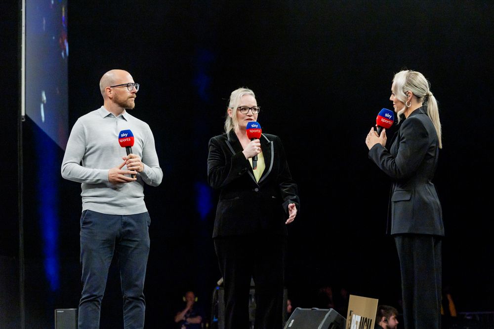 Three people, two men and one woman, stand on a stage and speak into microphones.