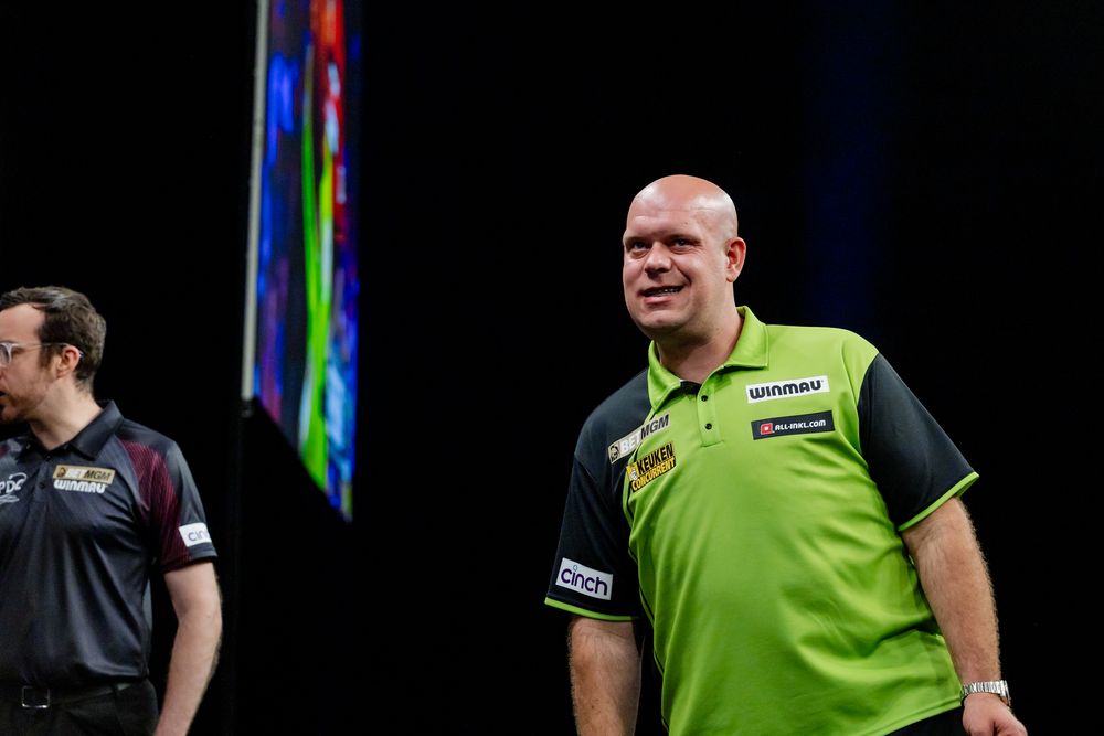A professional darts player in a green jersey standing on a stage during a darts tournament.