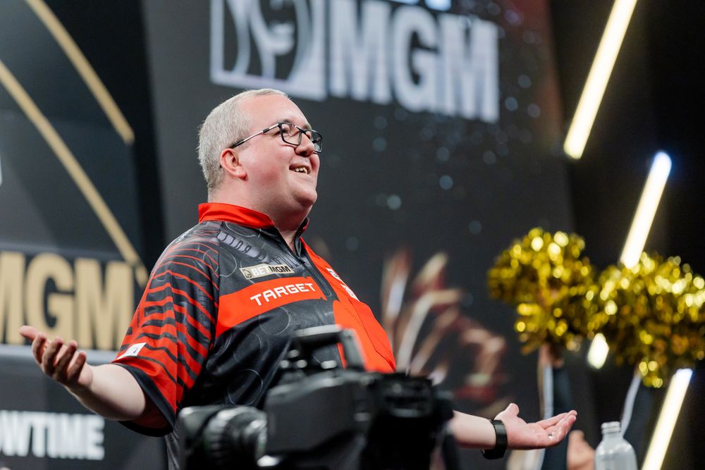 A professional darts player in a red and black jersey speaking on a stage during a darts tournament.