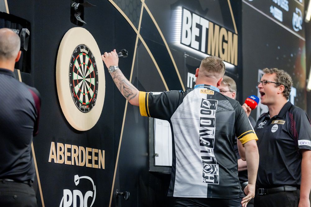 Two men are standing in front of a dart board, one appears to be throwing a dart.