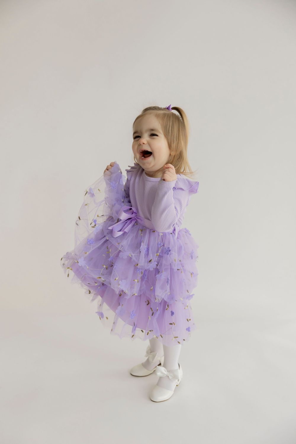 A toddler in a purple tulle dress and white shoes twirls and dances playfully against a light background.