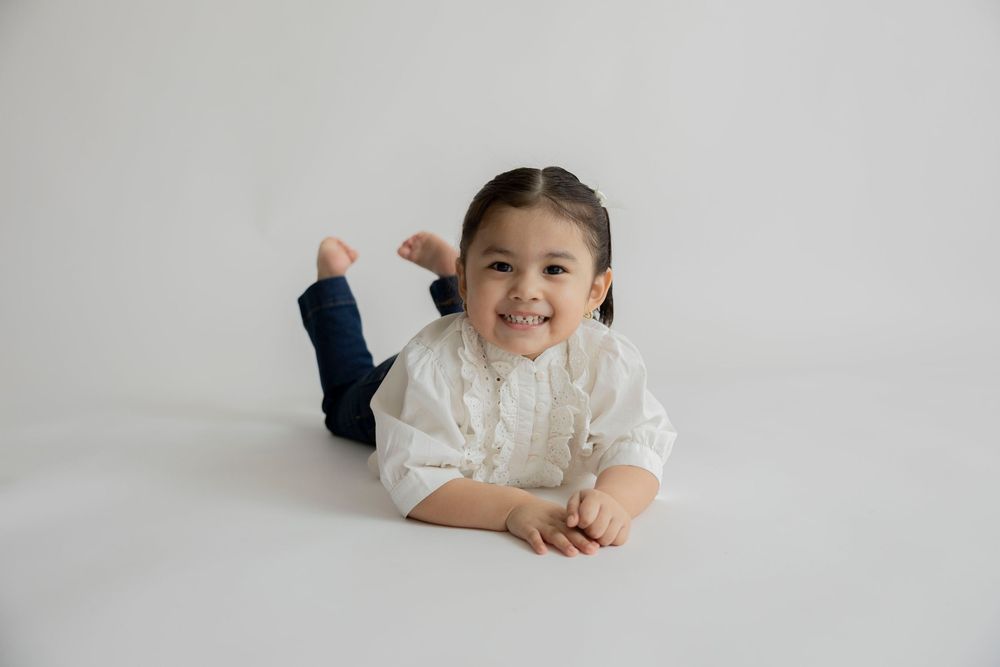 A child lies on a white background wearing a white sweater while smiling and playing.