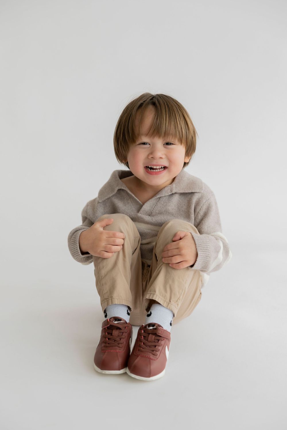 A young child in a beige outfit and brown shoes sits and smiles against a white background in a series of candid poses.