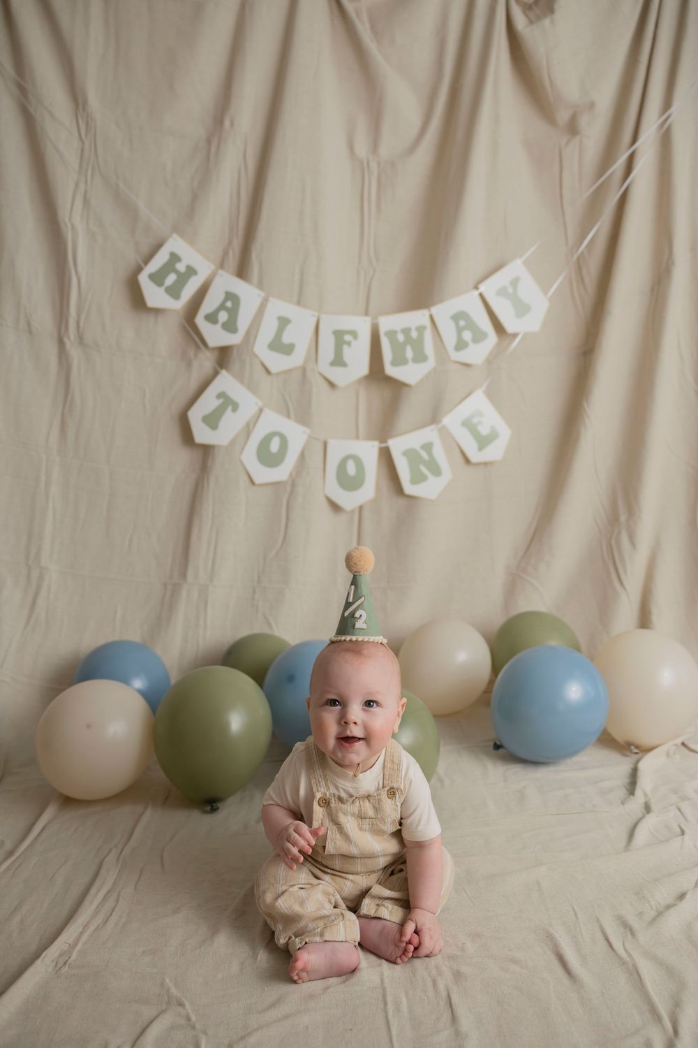 A baby in tan romper sits on cream backdrop with blue and green balloons and "halfway to one" banner for milestone photos in his Lake Orion