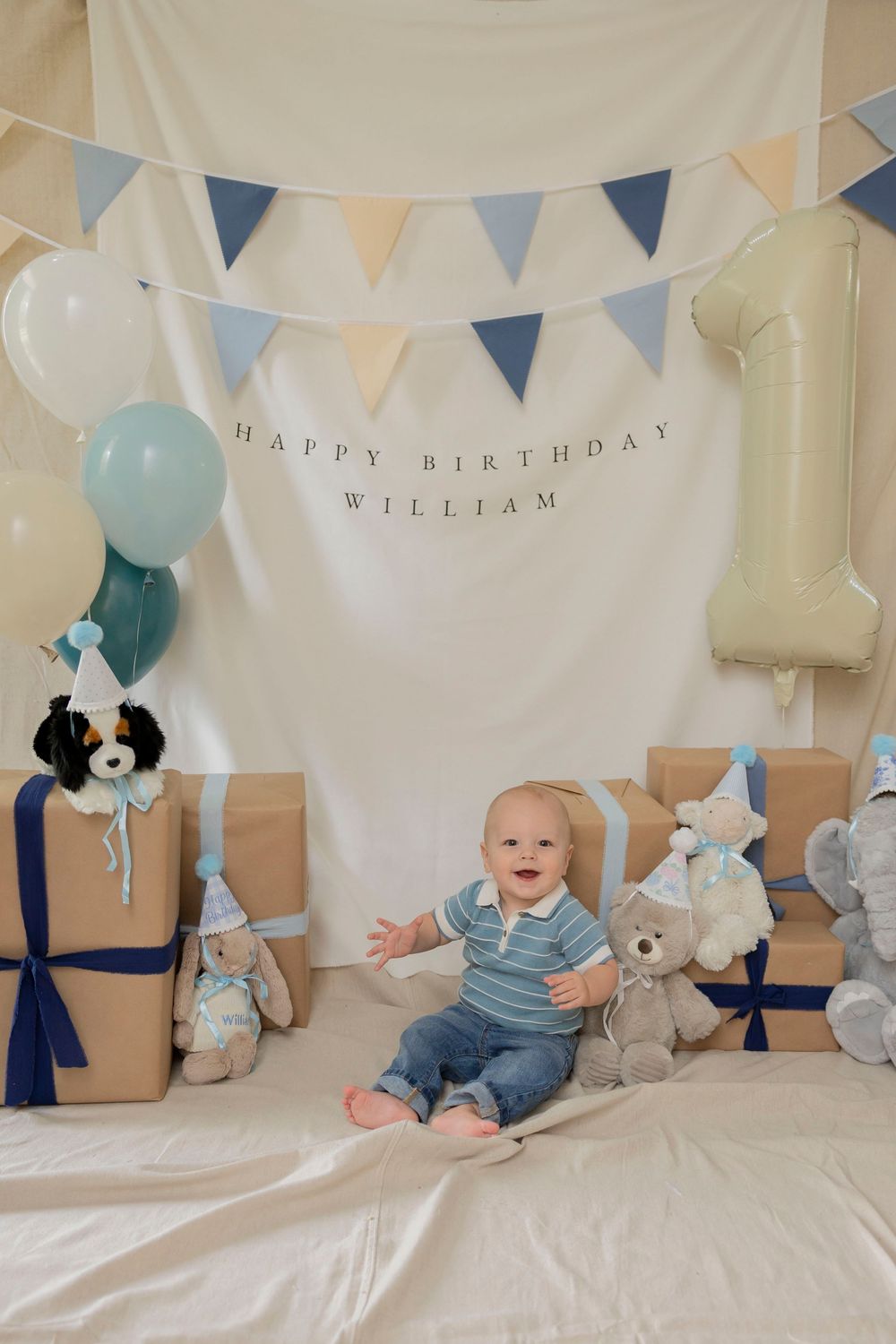 Baby sits smiling among gifts and teddy bears at his first birthday party with blue bunting and balloons in his Lake Orion michigan home