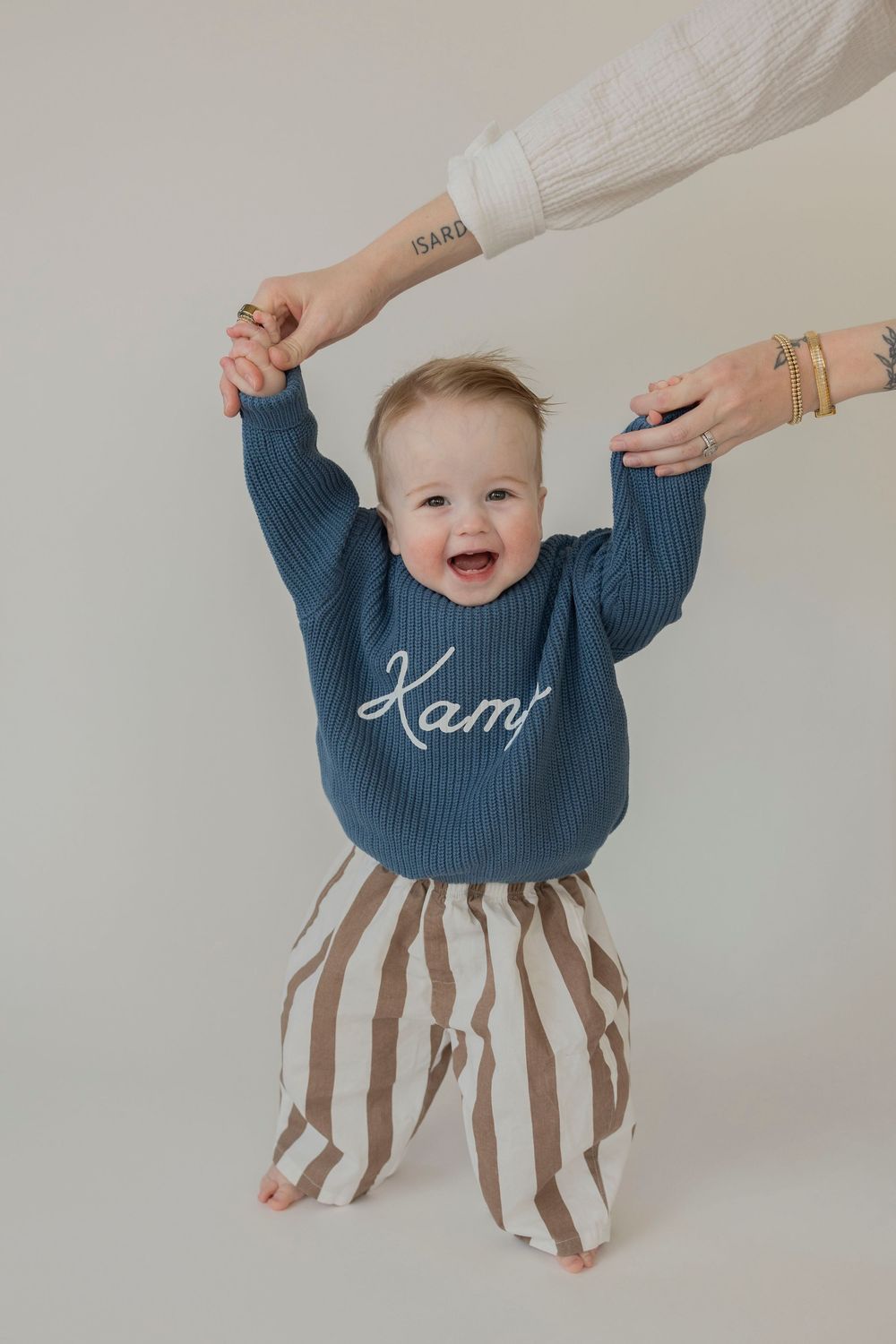 A baby in a navy sweater and striped pants holds hands with adults while standing against a light background for a 1 year old photoshoot