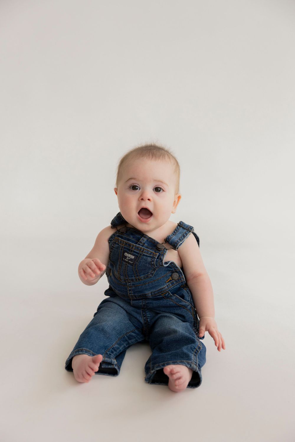 A baby wearing denim overalls sits and smiles on a white backdrop in a series of adorable studio portraits