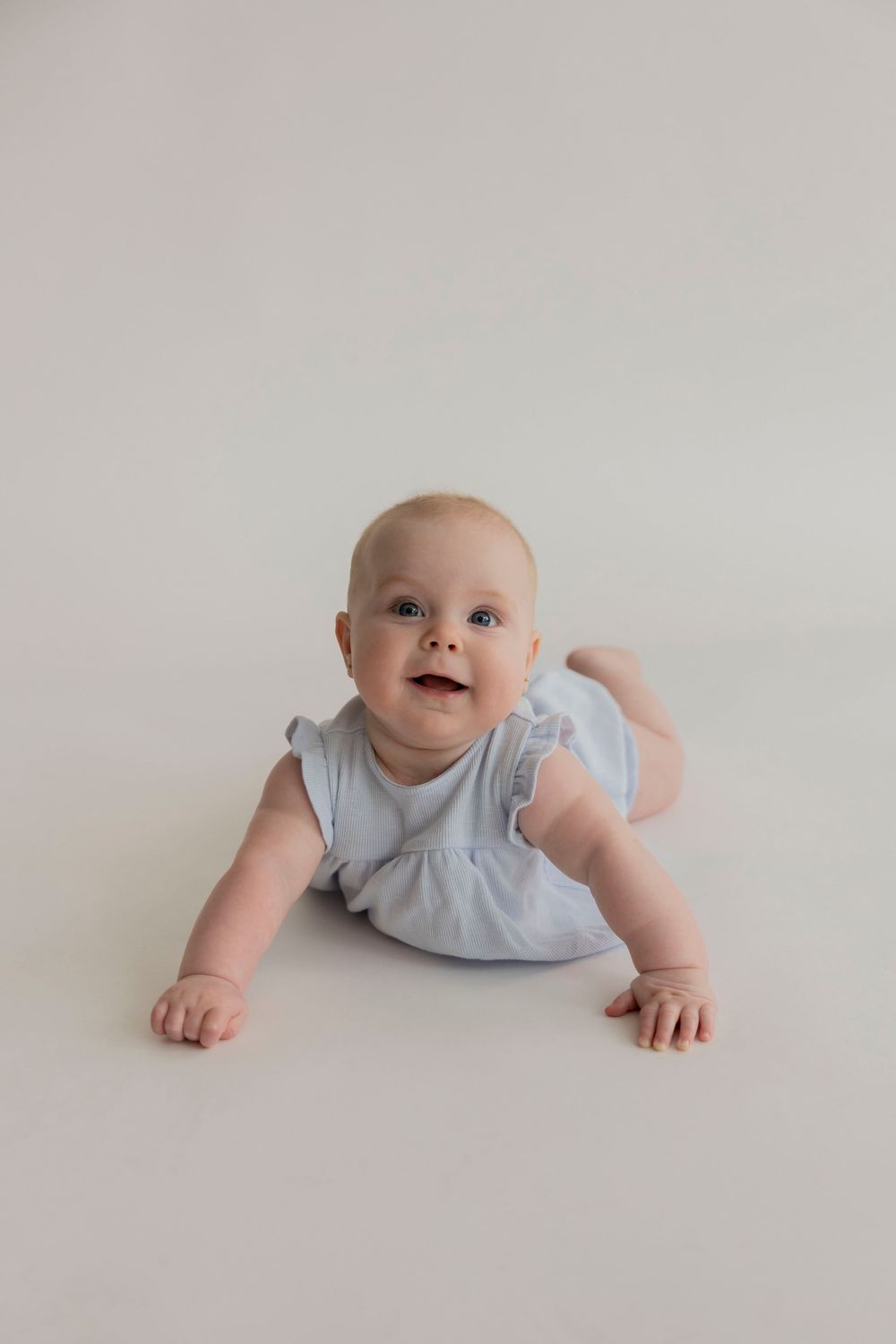 A baby in a light blue outfit crawls on a white background for a milestone photoshoot in her royal oak home