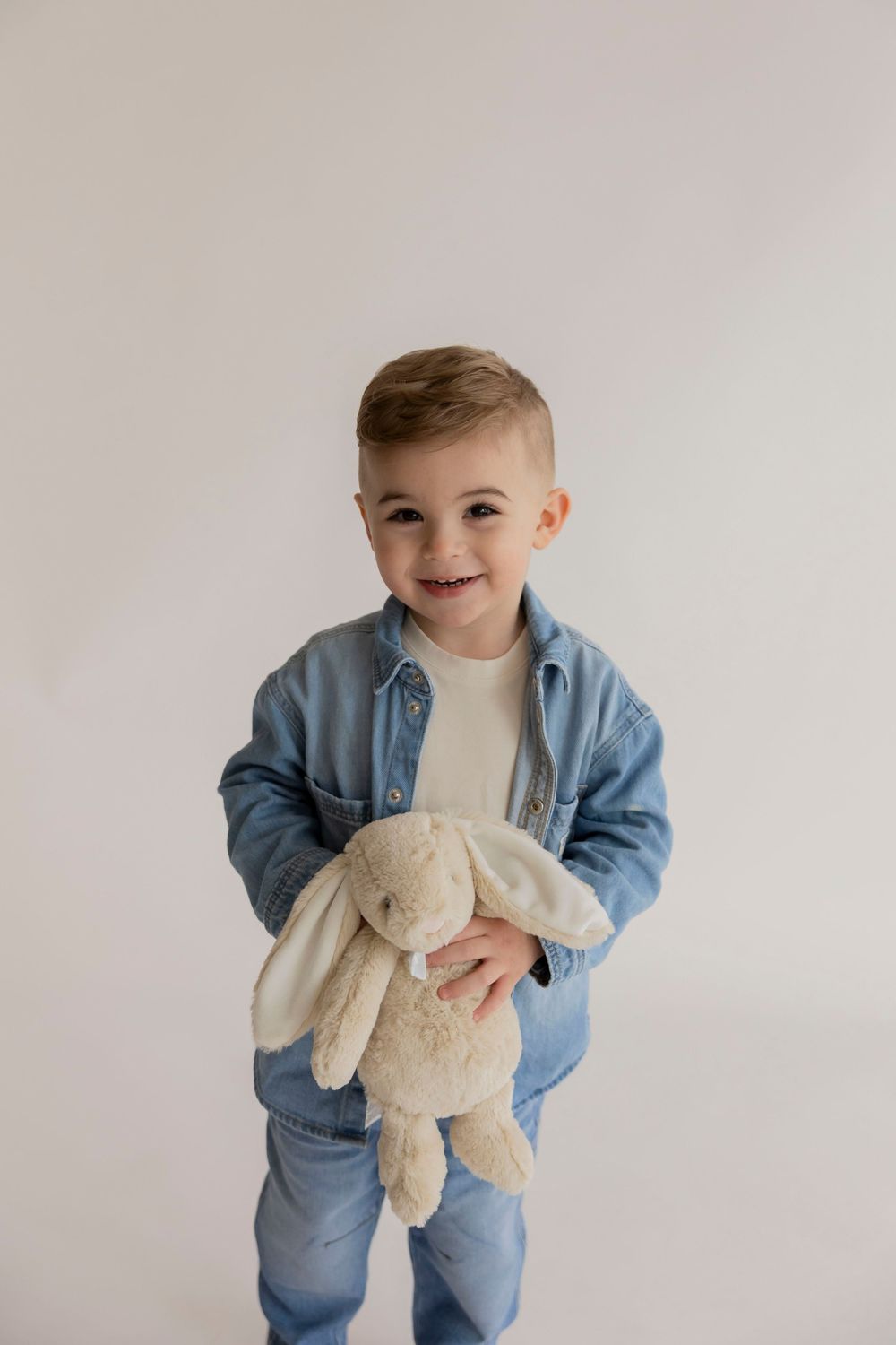 A small child in a blue denim jacket holds a plush bunny toy against a white background for 2 year old photos in his Rochester hills home