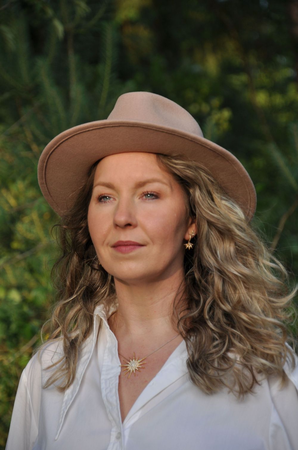 Close-up portrait of blonde woman in tan wide-brim hat with sun earrings and necklace outdoors.