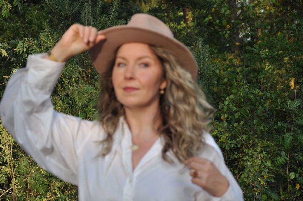 Smiling blonde woman in white shirt adjusting tan fedora hat with greenery background.