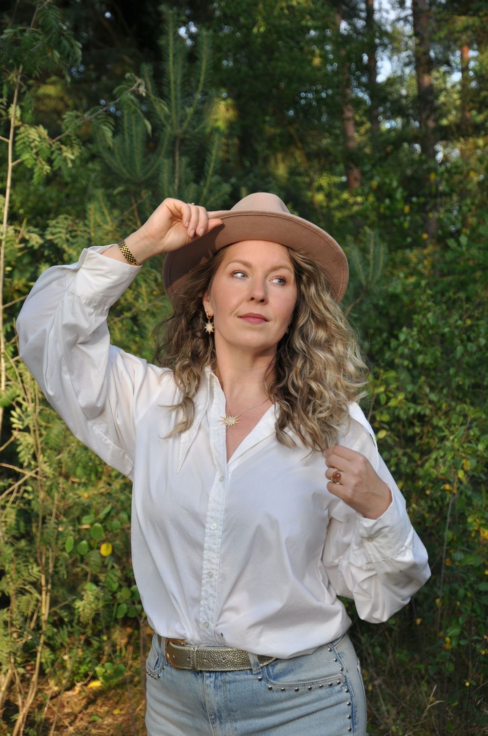 Woman in white blouse and jeans wearing a tan wide-brim hat posing in forest setting.