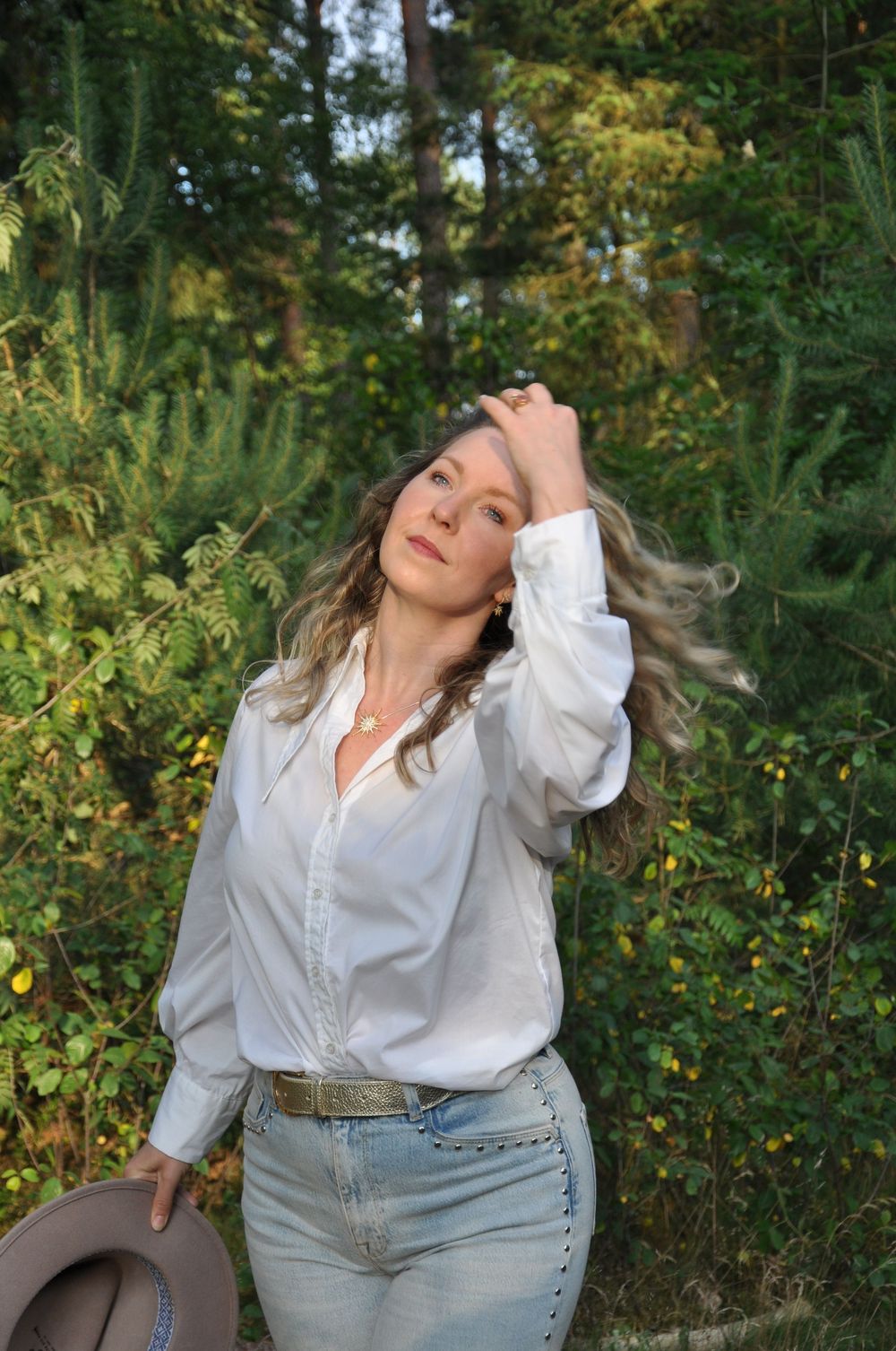 Blonde curly-haired woman in white blouse and jeans posing with hand in hair outdoors.