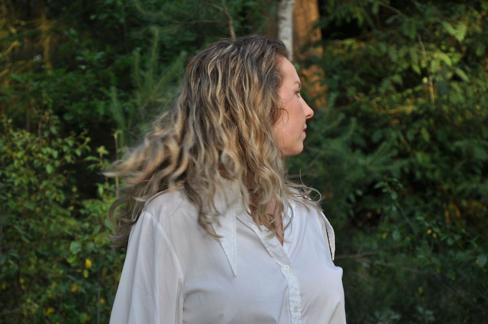 Side profile of curly-haired woman in white shirt gazing into green forest scenery.
