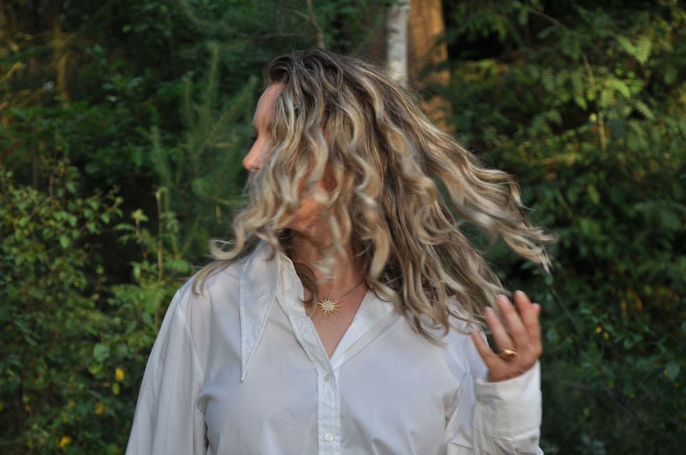 Woman with long curly blonde hair tossing her hair against a lush green forest background.