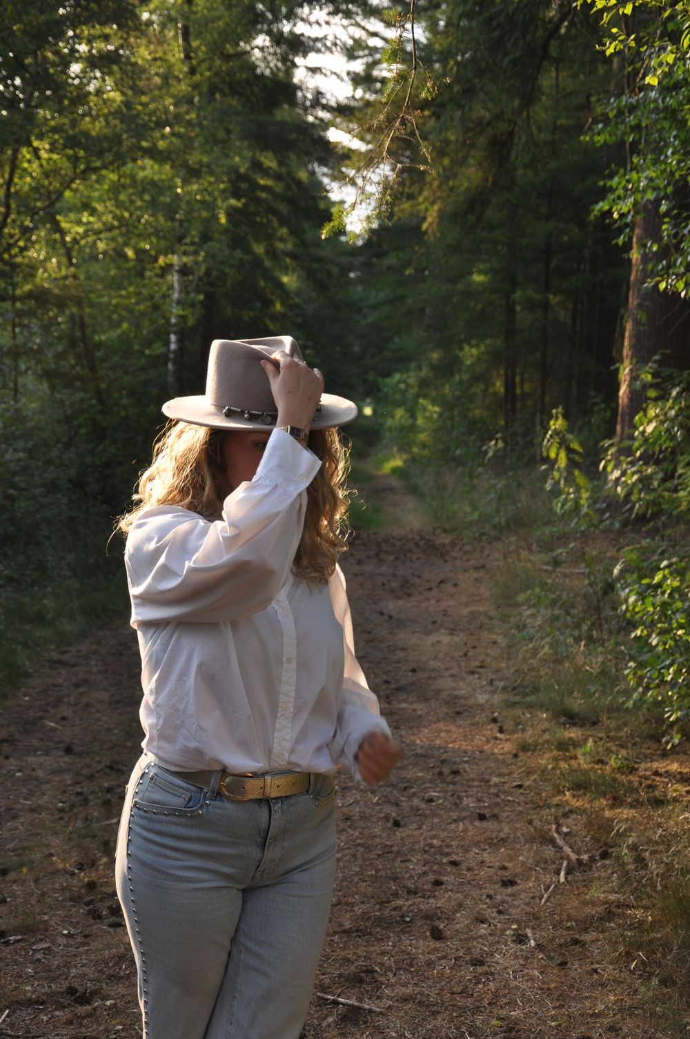 Woman in white blouse and jeans adjusting tan wide-brim hat on forest trail in golden light.