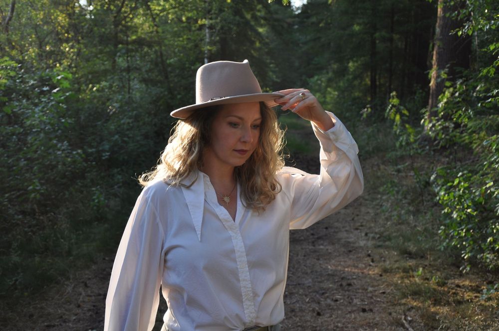 Blonde woman in white shirt tipping a tan fedora hat while standing on a woodland path.