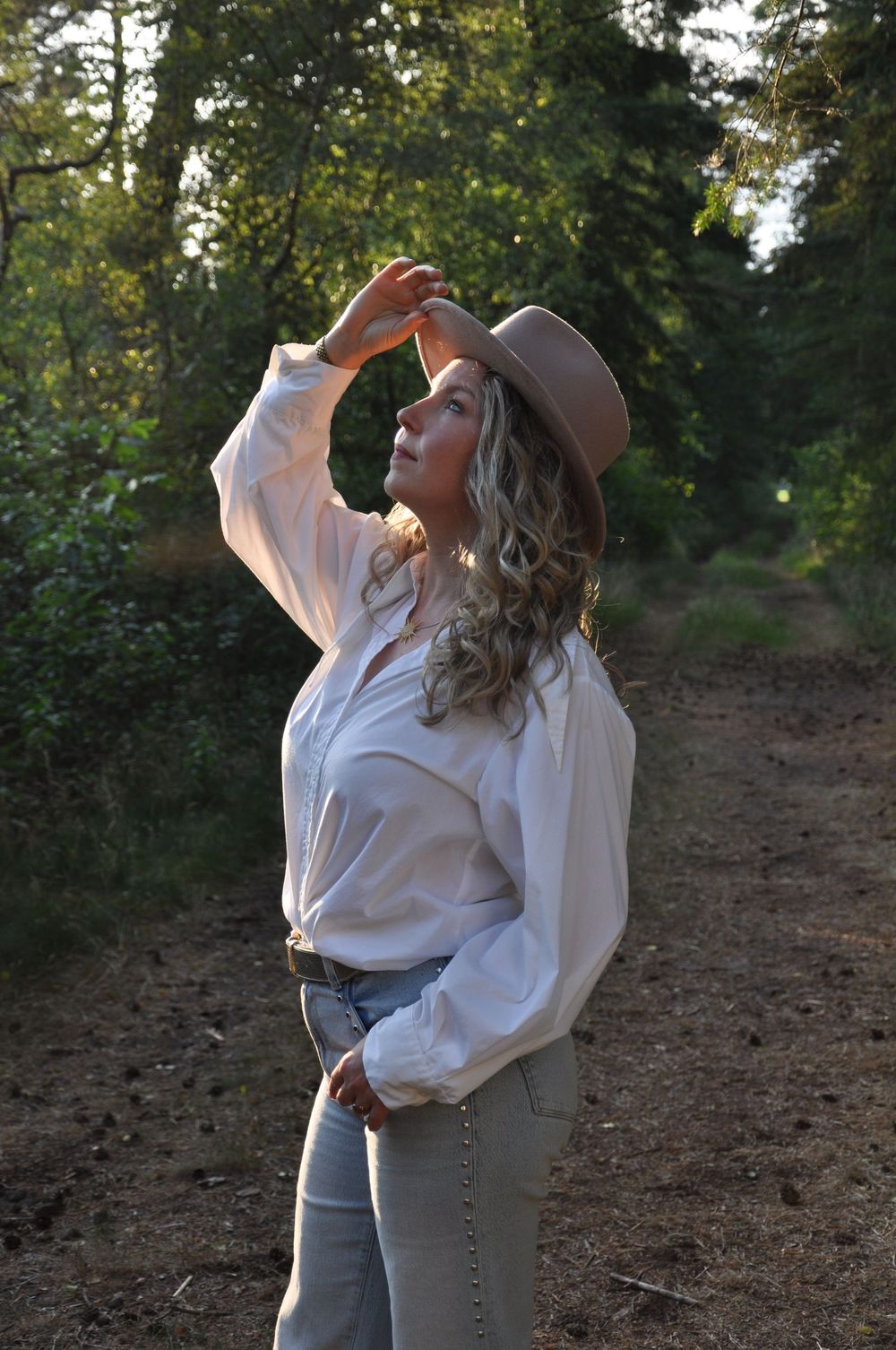 Blonde woman in white blouse and tan fedora hat standing on sunlit forest trail.