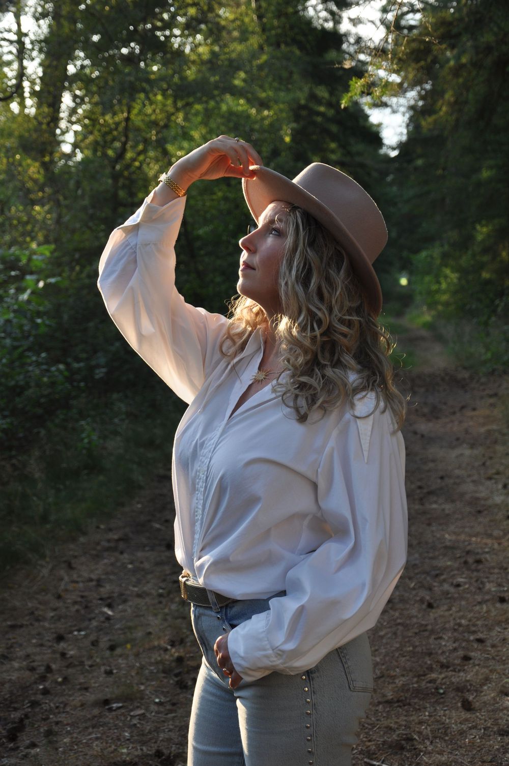 Blonde curly-haired woman in white blouse and tan hat posing in forest path.